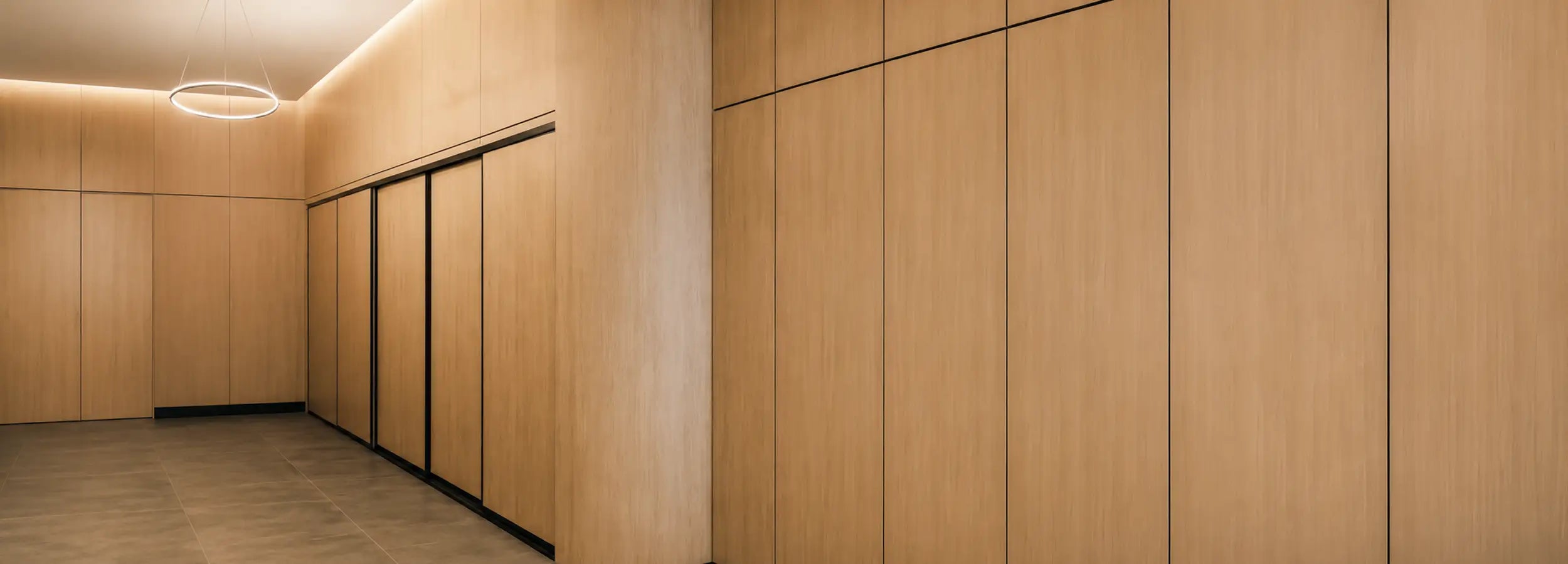 A modern interior space with light wood paneling on the walls and a matching wood floor, featuring a row of sliding doors with slim black handles and a circular ceiling light.
