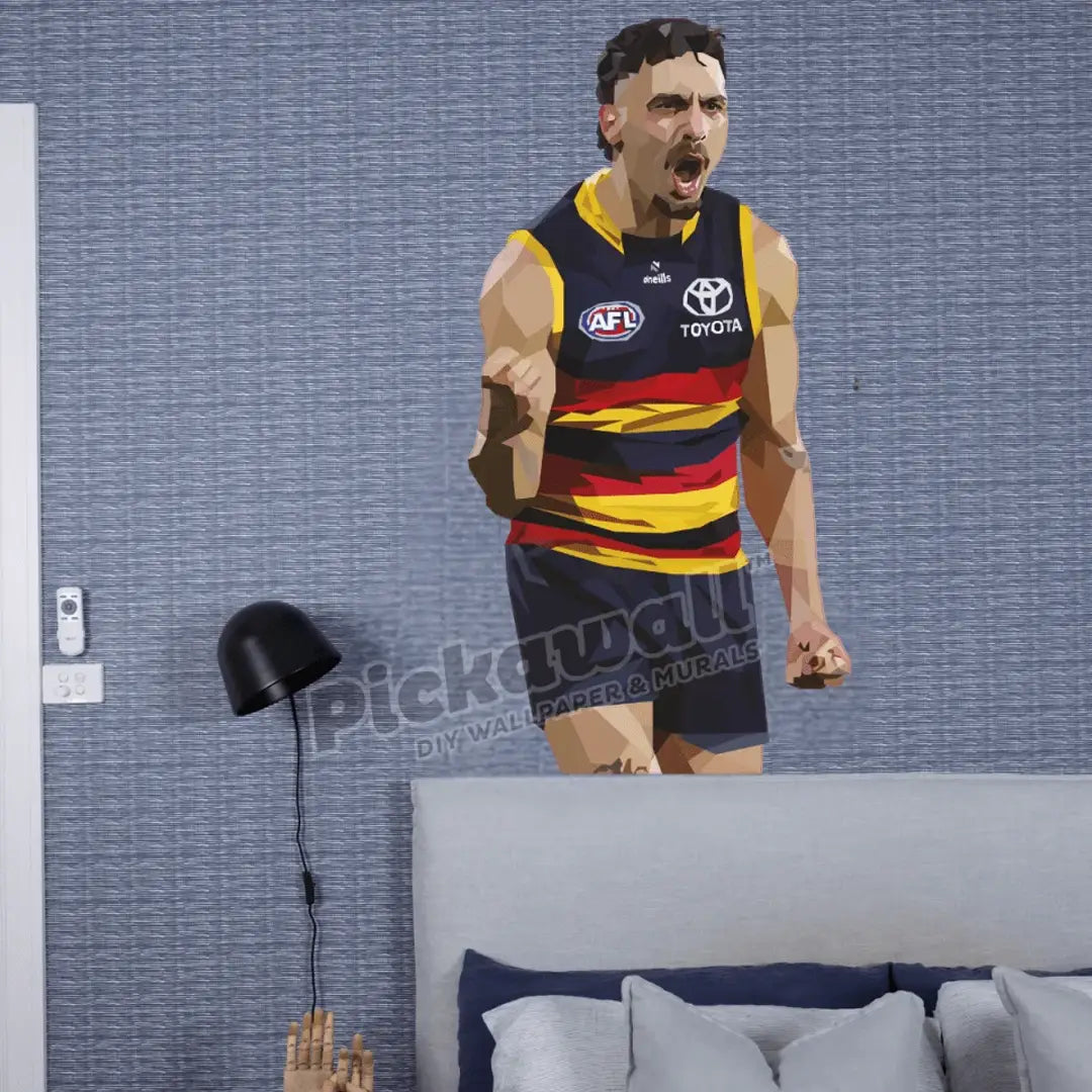 A large wall decal of an AFL player in a yellow, red, and navy striped jersey is shown applied to a textured blue-grey wall above a bed, with a black floor lamp on the left. A 'Pickawall DIY WALLPAPER & MURALS' watermark is visible across the centre.